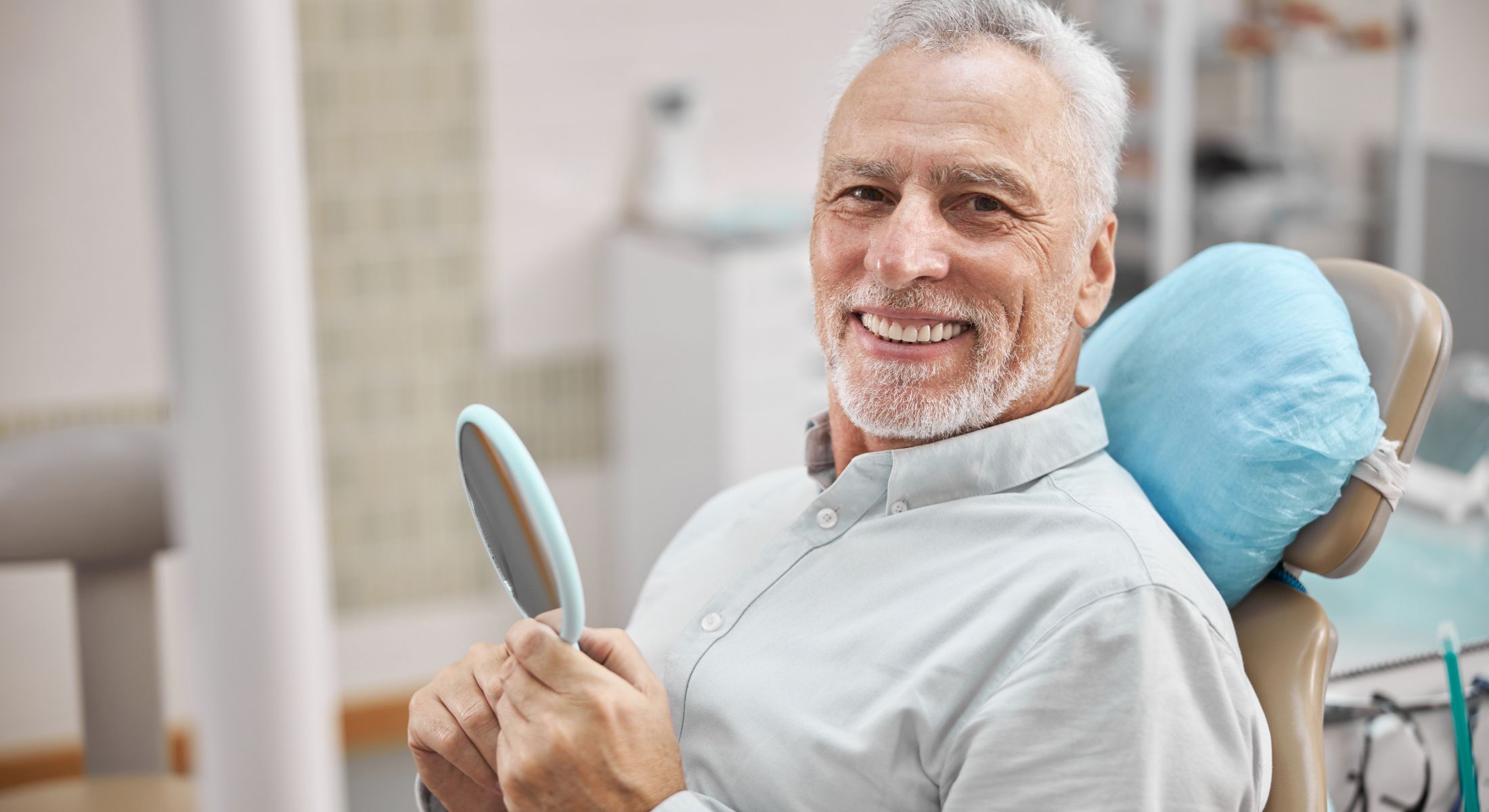 Smiling elderly man in dental chair holding mirror.